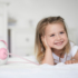 Kép 6/11 - Mosolygós kislány az ágyon könyököl, mellette Pink Nosiboo pro2 elektromos orrszívó